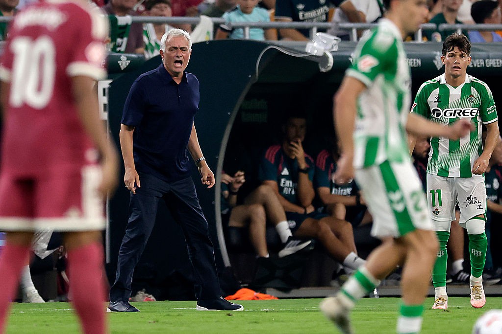 O técnico José Mourinho, de cabelos grisalhos e camisa polo azul-marinho, grita com a boca aberta na beira do campo, enquanto jogadores em uniformes de futebol verde e branco e vermelho desfocam em primeiro plano. Ao fundo, jogadores reservas sentam no banco