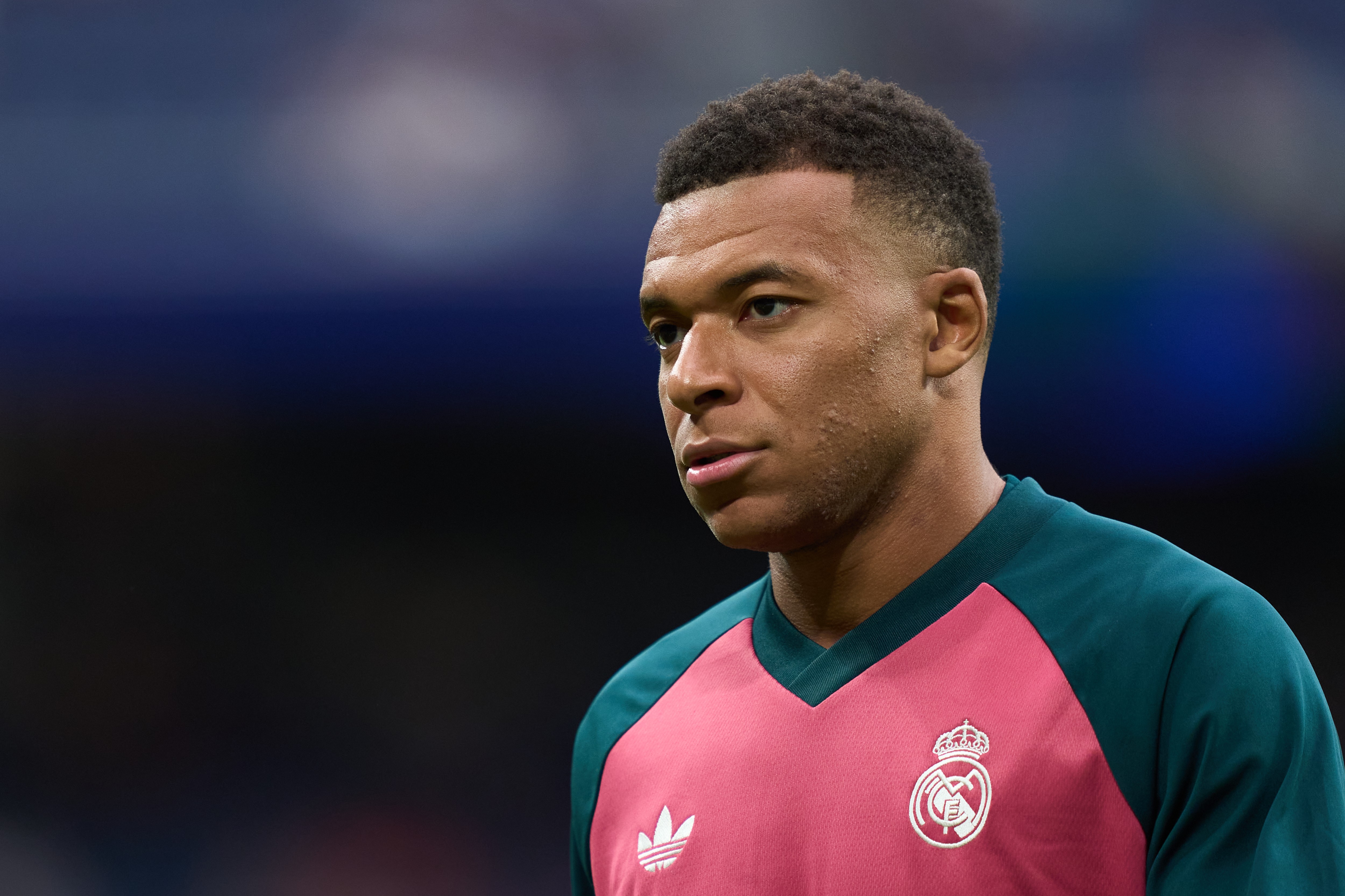 Kylian Mbappé, jogador de futebol, com corte de cabelo degradê, vestindo uma camisa de treino rosa com detalhes verdes e o escudo do Real Madrid, em um fundo escuro e desfocado de um estádio