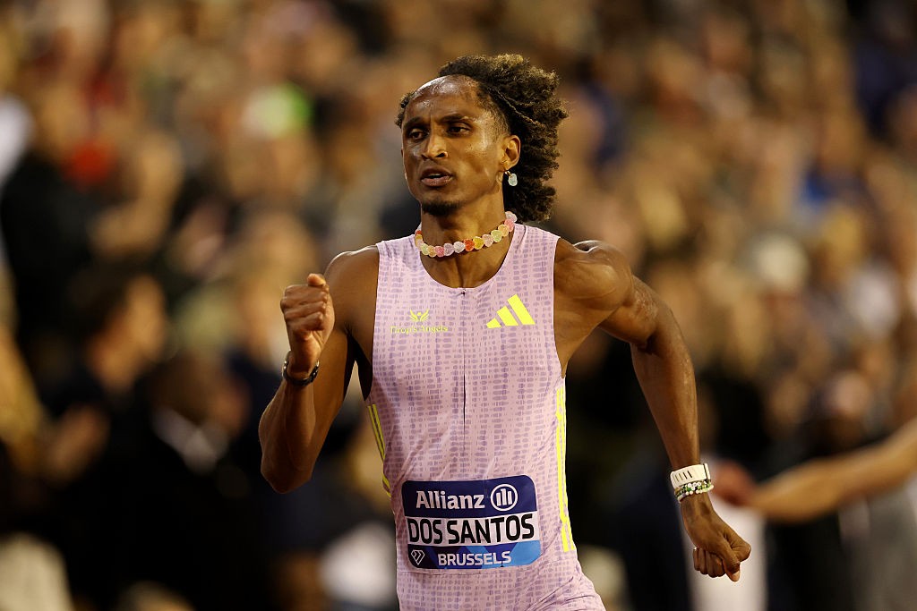 Alison dos Santos, atleta de pele escura e cabelo crespo, corre em pista de atletismo. Ele veste um macacão lilás com detalhes amarelos e um colar colorido. A expressão é concentrada, e o fundo está desfocado com a plateia. No peito, um número de identificação com Allianz DOS SANTOS BRUSSELS