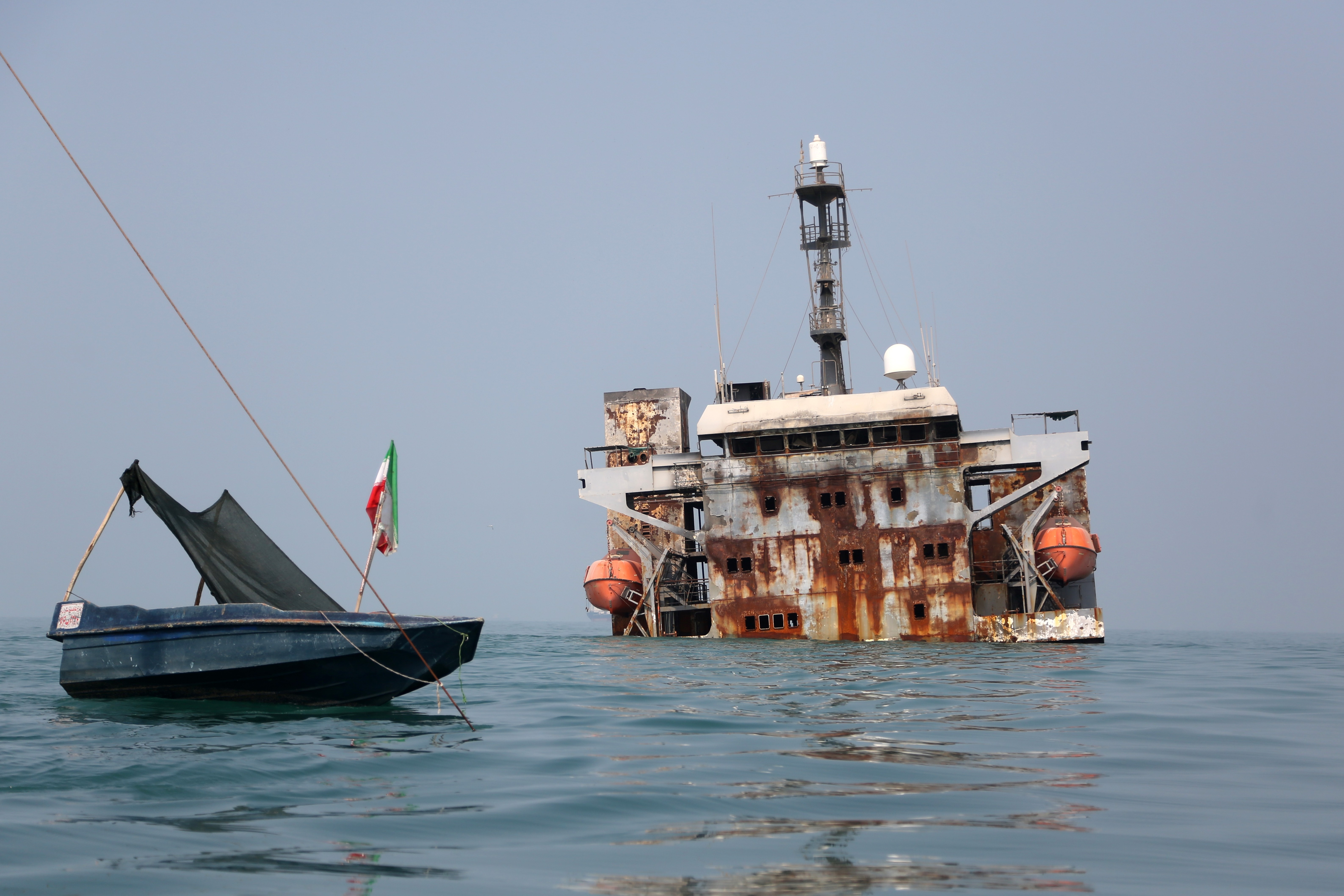 Um pequeno barco azul com uma bandeira iraniana e uma lona preta, amarrado por uma corda, flutua em águas calmas. Ao fundo, um navio enferrujado e parcialmente submerso, com botes salva-vidas laranja e uma torre de comunicação, emerge da água sob um céu claro.