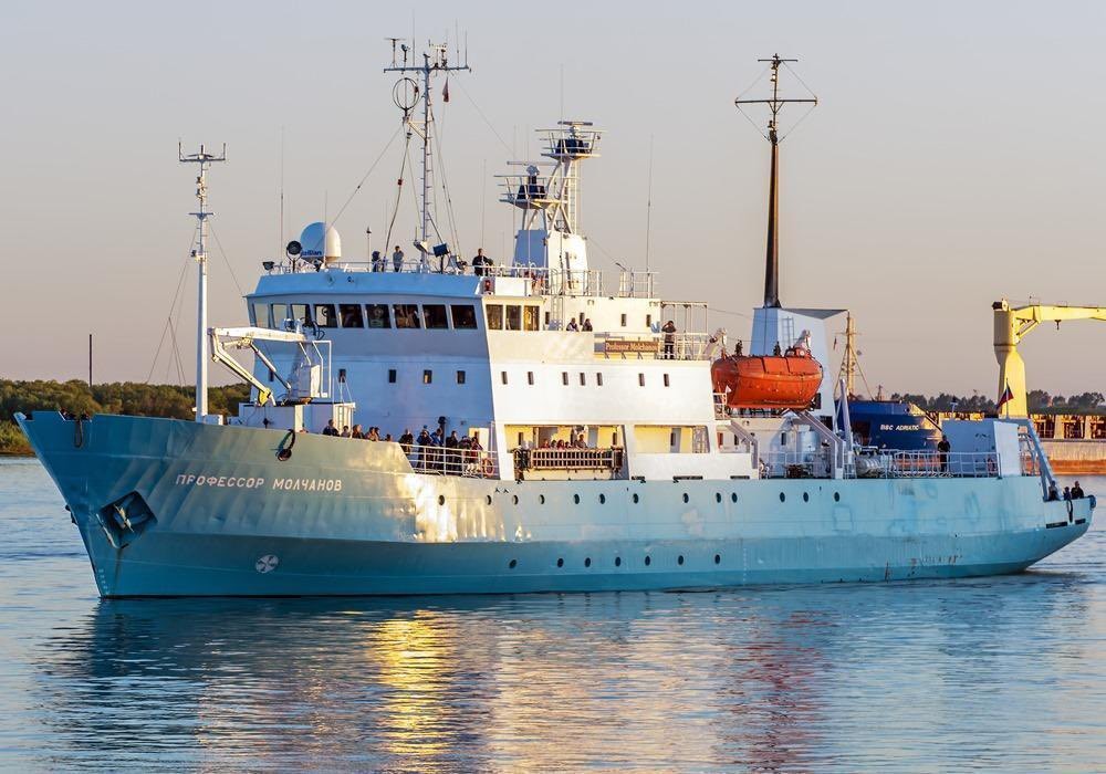 Navio de pesquisa Professor Molchanov, de cor azul-claro e branco, navegando em águas calmas sob um céu claro. Pessoas são visíveis nos conveses e varandas, e um bote salva-vidas laranja está pendurado na lateral. Guindastes e antenas se destacam no topo da embarcação, com reflexos dourados do sol na água