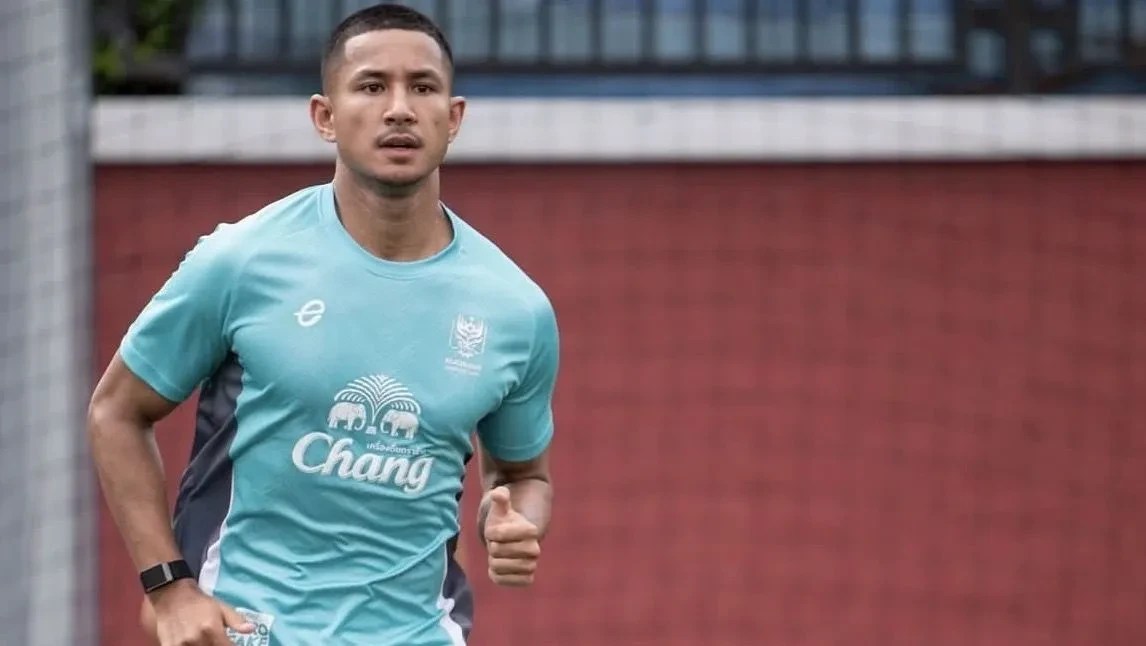 Homem asiático com cabelo curto e barba rala, vestindo camisa de treino azul-clara com logo de elefantes e a palavra Chang, correndo com expressão séria, em frente a uma parede de tijolos vermelhos