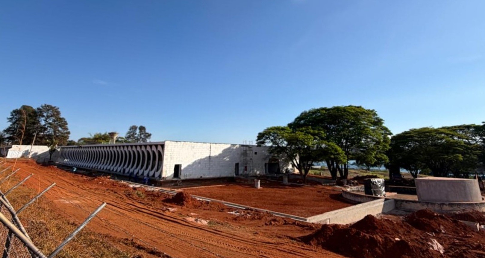 Construção de um edifício branco longo e baixo com aberturas arqueadas, cercado por terra vermelha e árvores frondosas sob um céu azul claro