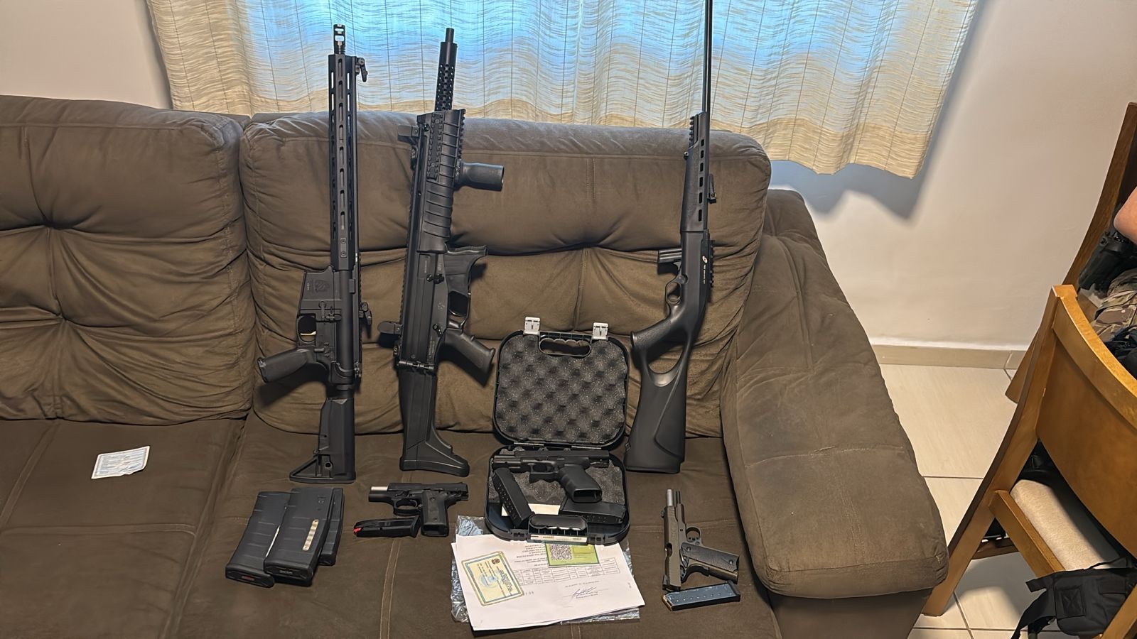 Armas de fogo apreendidas sobre um sofá marrom: três fuzis, duas pistolas, um revólver, carregadores e documentos em uma mesa de centro