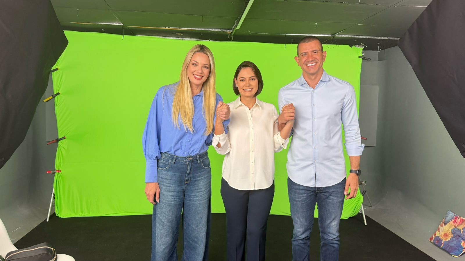 Três pessoas sorridentes, duas mulheres e um homem, de mãos dadas em frente a um fundo verde de chroma key. A mulher à esquerda tem cabelos loiros longos e veste camisa azul e jeans. A mulher central tem cabelos curtos escuros e veste camisa branca e calça escura. O homem à direita veste camisa azul clara e jeans.