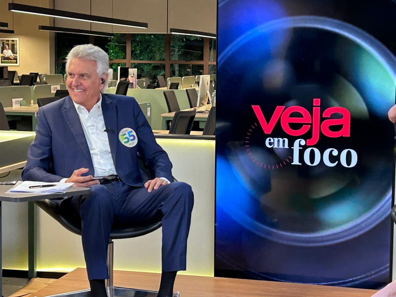Homem de cabelos grisalhos, terno azul e camisa branca, sorri sentado em uma cadeira preta, com um broche 55 na lapela. À direita, uma tela exibe o logo Veja em Foco em vermelho e branco sobre fundo escuro com anéis azuis