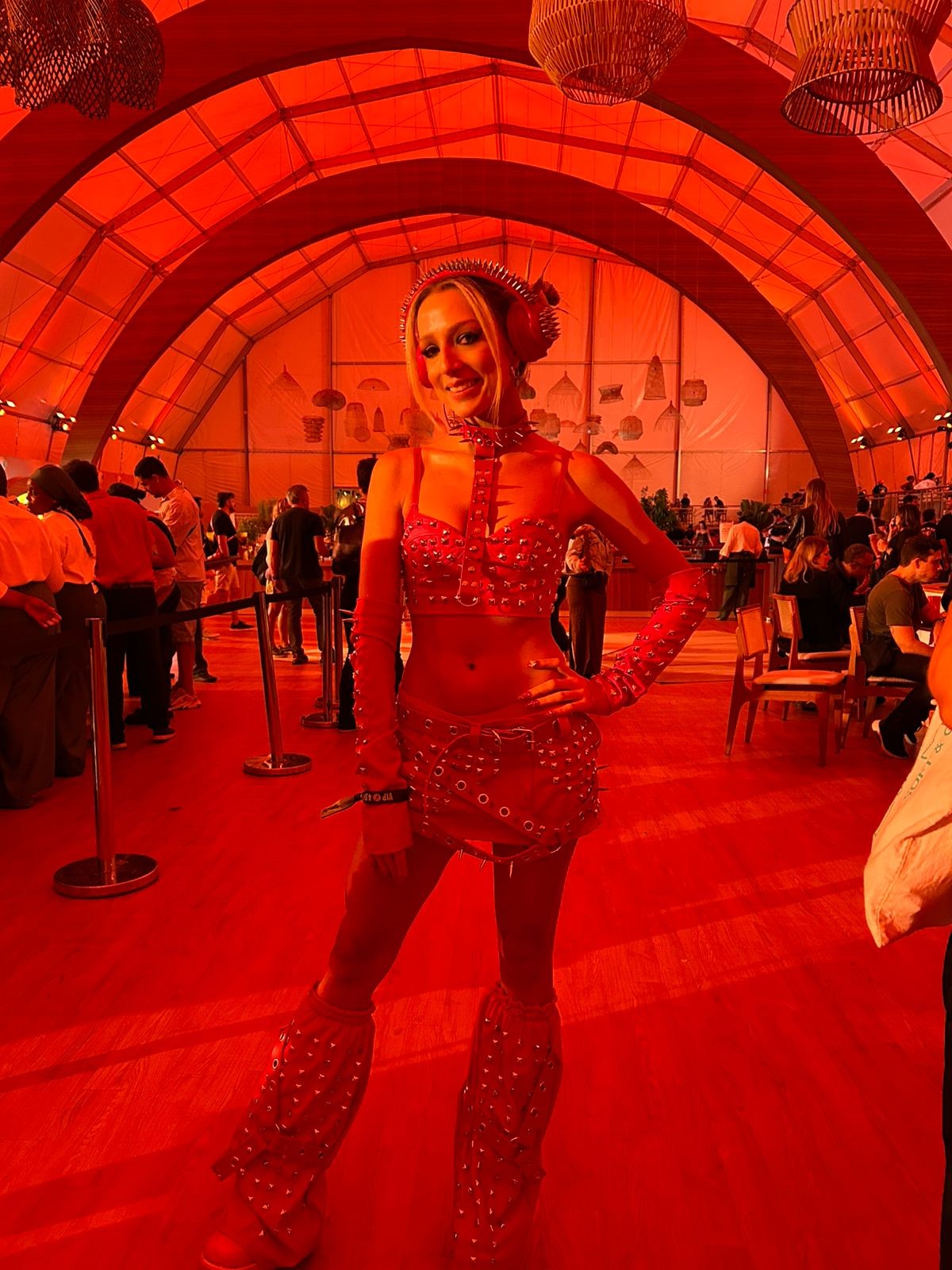 Mulher loira sorrindo, vestindo top, minissaia e botas vermelhas cravejadas, com fones de ouvido e gargantilha de spikes, em um evento com iluminação vermelha e teto abobadado