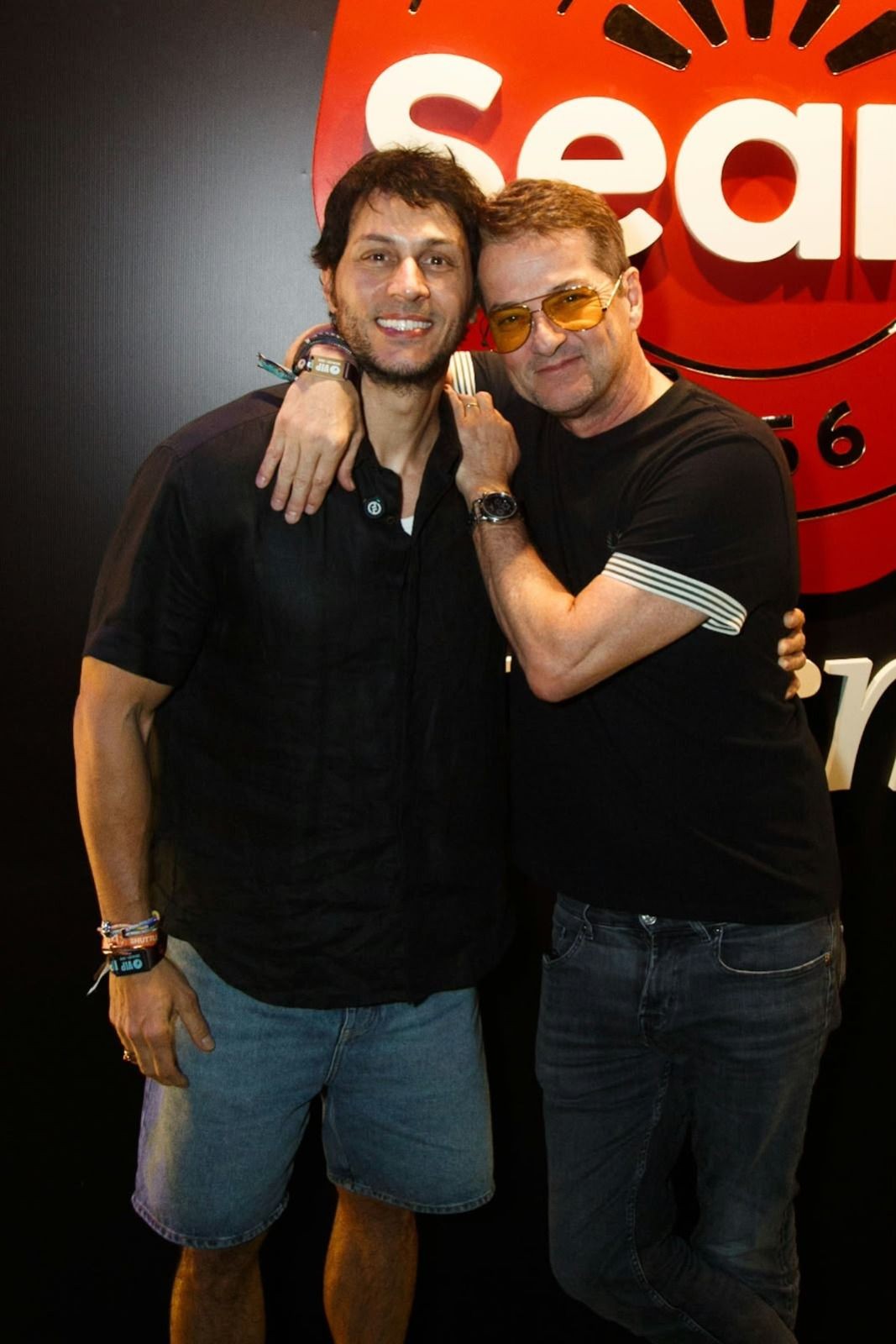 Dois homens sorridentes posam abraçados, o da esquerda veste camisa preta e bermuda jeans, o da direita usa óculos de sol amarelos, camiseta preta com listras brancas na manga e calça jeans escura, em frente a um letreiro vermelho com a palavra Seara