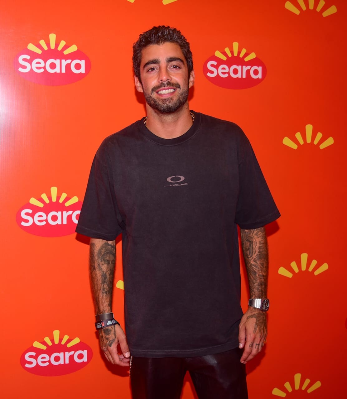 Homem branco, barba e cabelo escuros, sorrindo para a câmera, vestindo camiseta preta e calça de couro. Ele exibe tatuagens nos braços e um relógio prateado no pulso direito. O fundo é laranja com logotipos da Seara