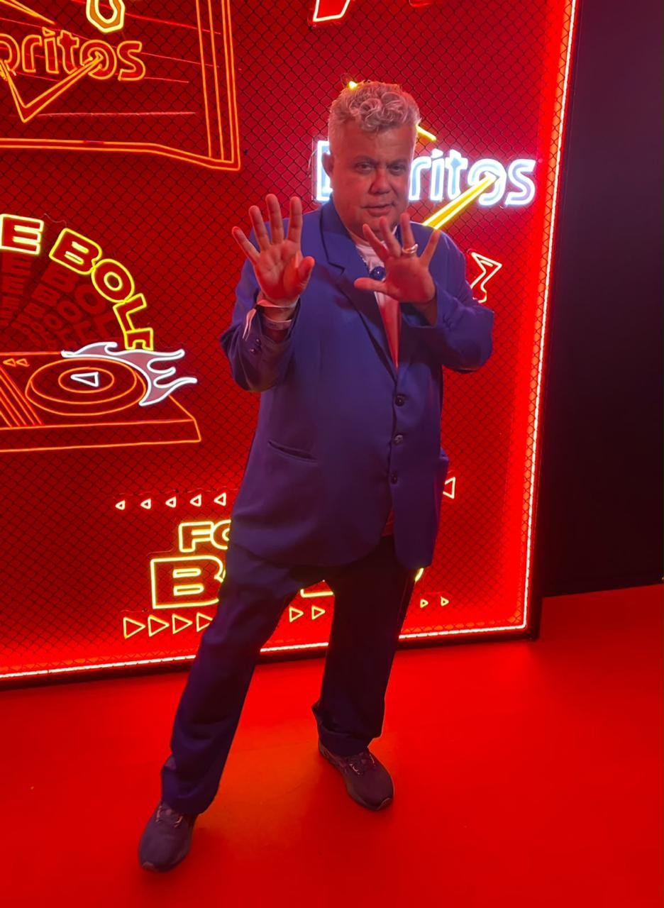 Homem de cabelos grisalhos e encaracolados, vestindo blazer azul e camiseta rosa, posa com as mãos abertas para a câmera. Ele está em um ambiente com iluminação vermelha e letreiros de neon com as palavras Doritos e Ebola
