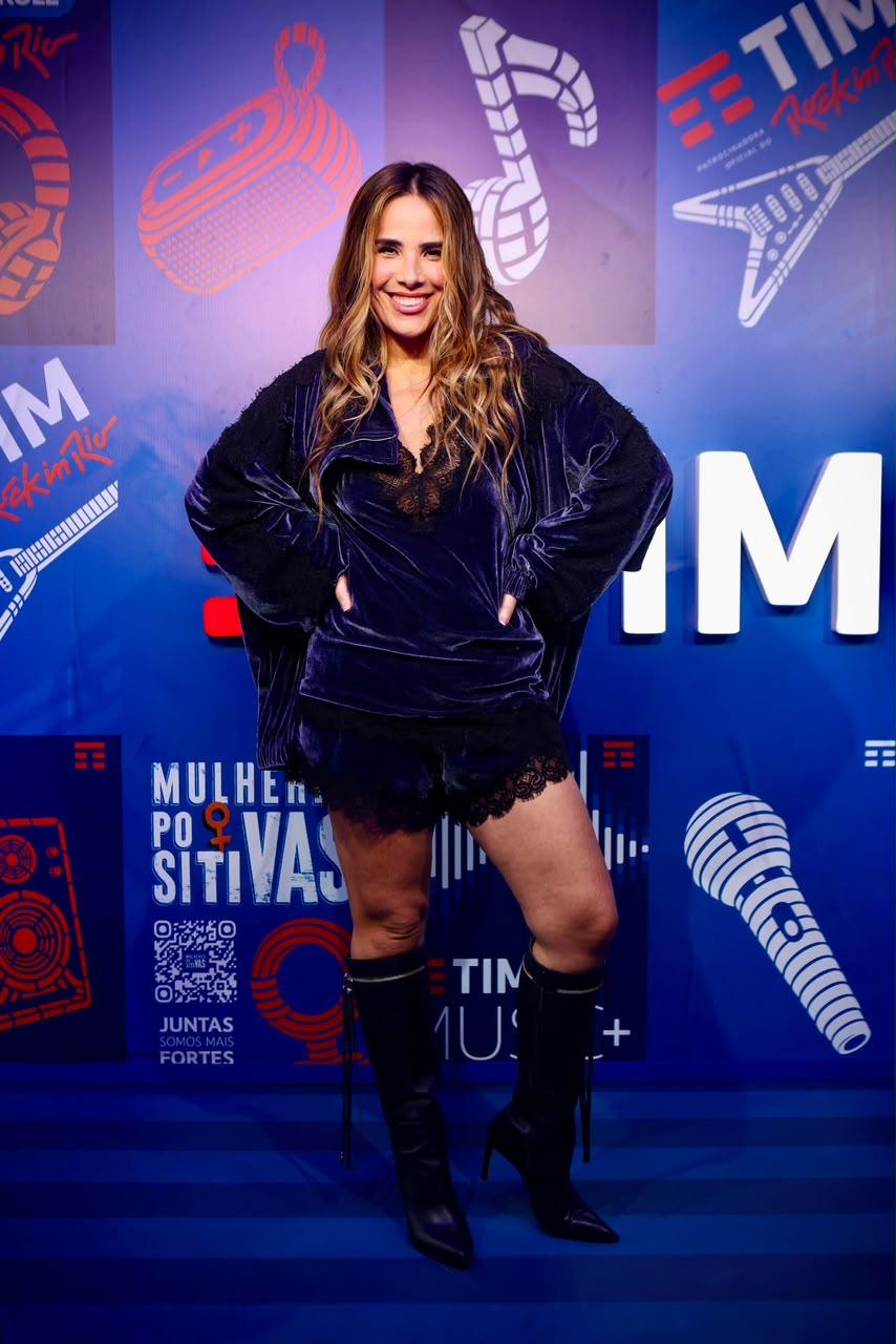 Mulher sorridente de cabelos longos e ondulados, vestindo conjunto de veludo azul-marinho com renda preta e botas de cano alto, posa com as mãos na cintura em frente a um painel azul com logotipos e ilustrações musicais