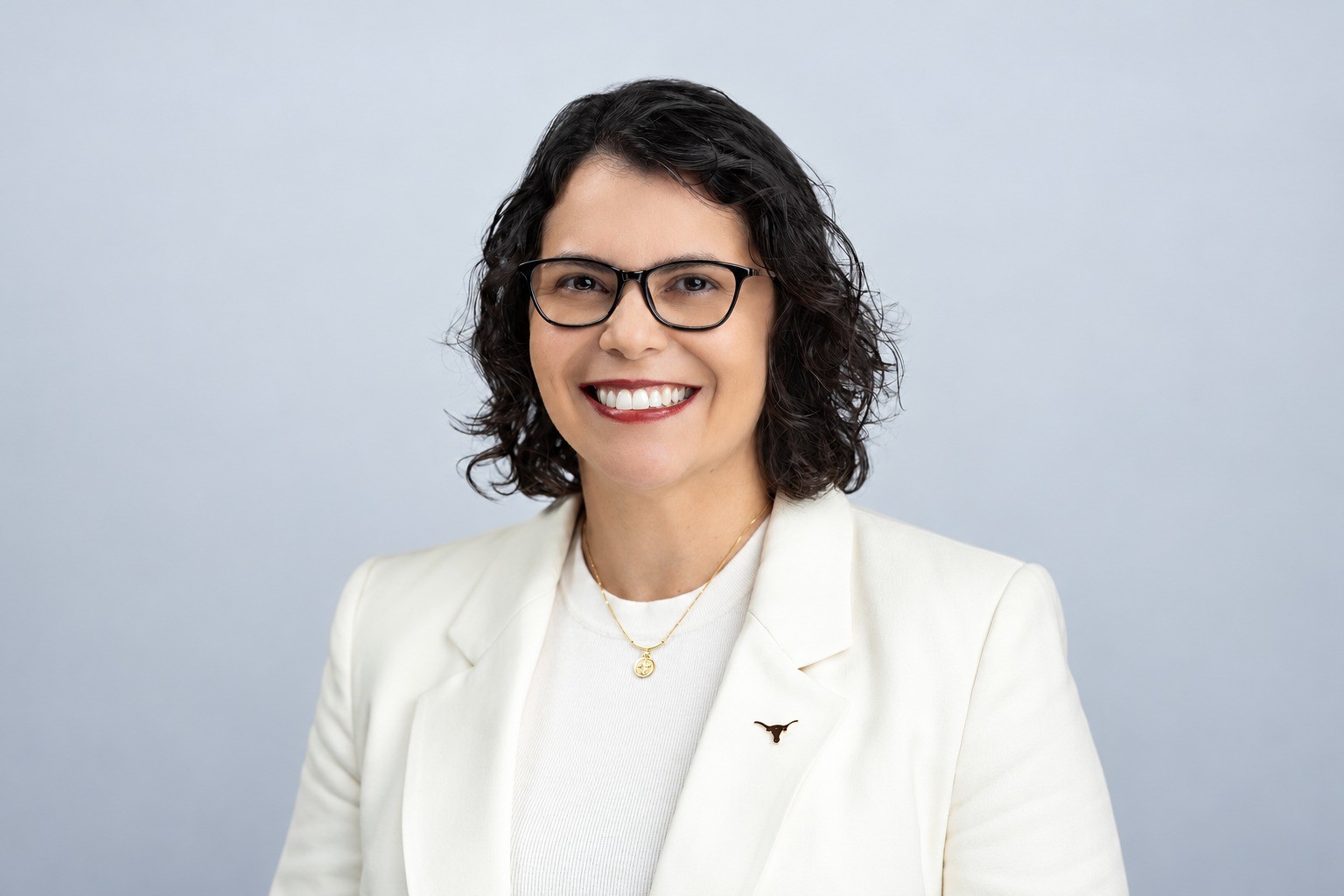 Mulher de pele clara, cabelos escuros e encaracolados, sorrindo, usando óculos pretos, blazer creme e colar dourado, em fundo cinza claro