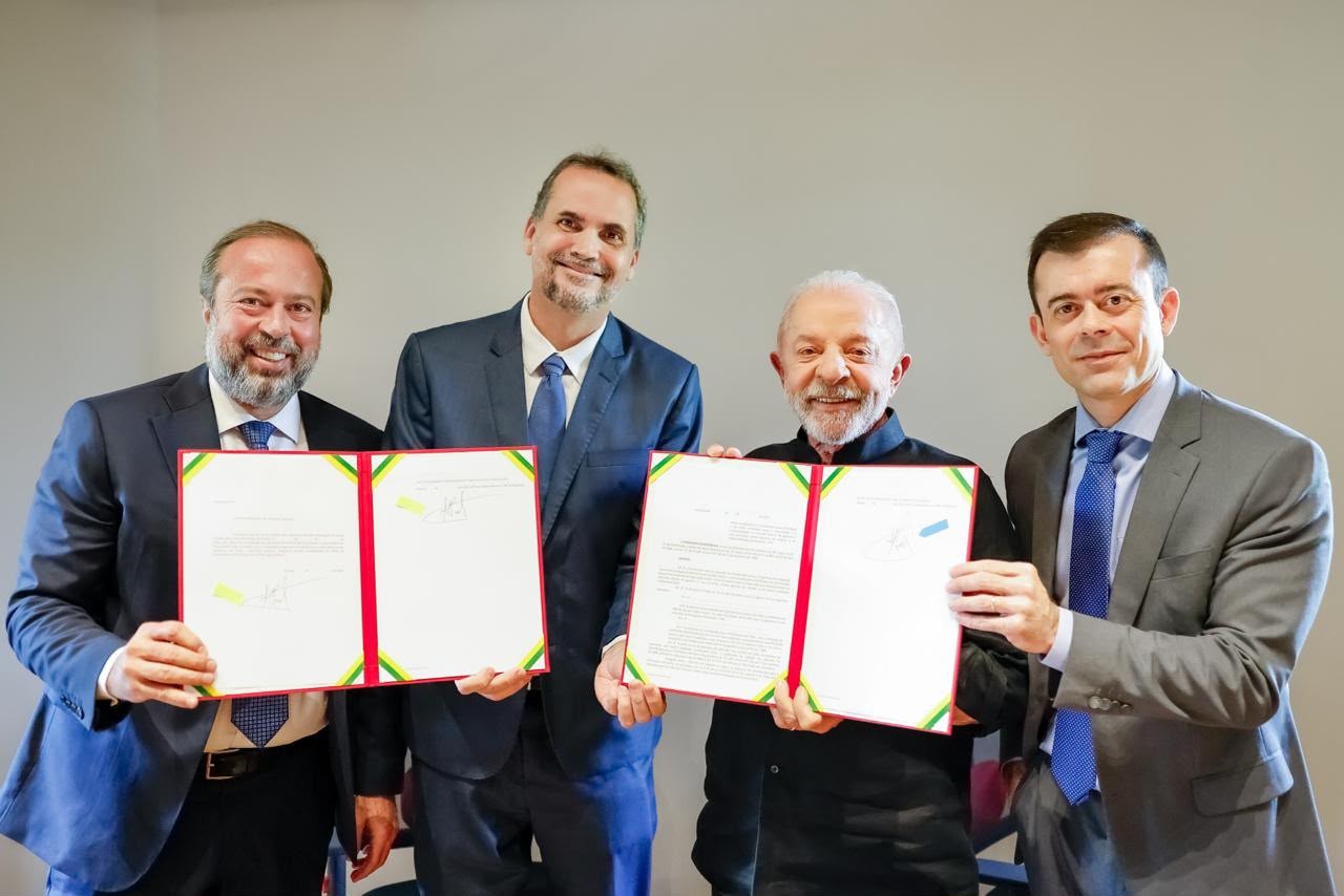 Quatro homens sorridentes, dois de terno e gravata e dois de terno, seguram documentos abertos com bordas em vermelho e amarelo, em um fundo cinza claro