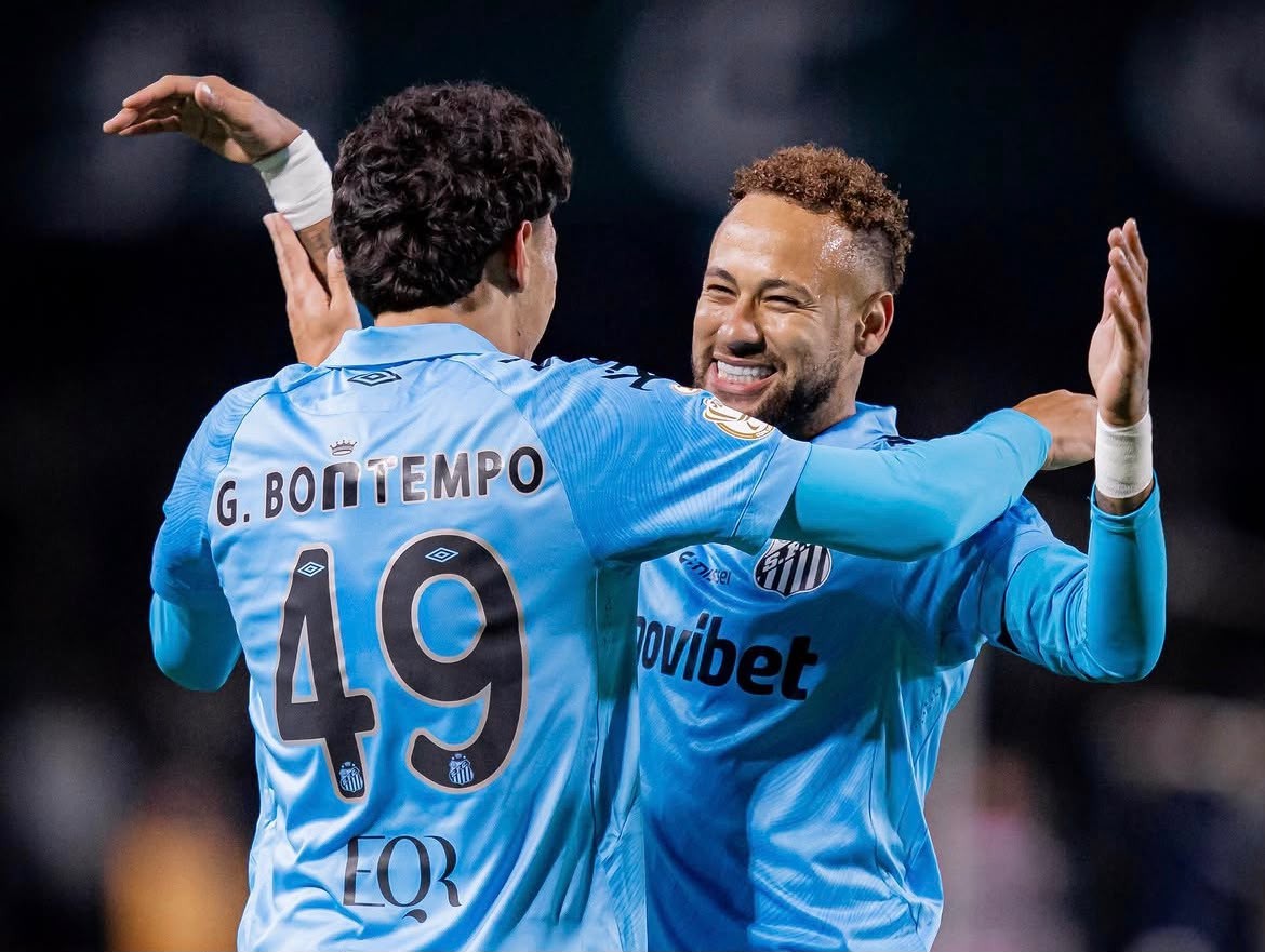 Dois jogadores de futebol, um de costas com a camisa 49 e outro de frente sorrindo, se abraçam em campo, ambos vestindo camisas azuis