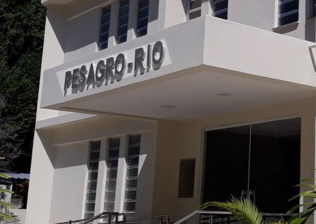 Fachada de um prédio branco com a inscrição PESAGRO-RIO em letras cinzas em relevo sobre uma faixa horizontal. Abaixo, uma porta de vidro escuro e janelas verticais. Plantas verdes aparecem no canto inferior direito e uma área arborizada no canto superior esquerdo
