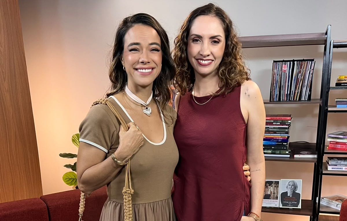 Duas mulheres sorridentes posam lado a lado em um ambiente interno. A da esquerda veste blusa marrom e saia plissada, com colar e bolsa de alça trançada. A da direita usa blusa regata vinho e colar discreto. Ao fundo, uma estante com livros e discos