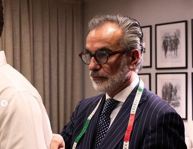 Um alfaiate de meia-idade, com barba e cabelos grisalhos, óculos escuros, terno azul-marinho risca de giz e gravata de bolinhas, mede um cliente de camisa branca com uma fita métrica nas cores da bandeira italiana. Ao fundo, cortinas e quadros emoldurados na parede