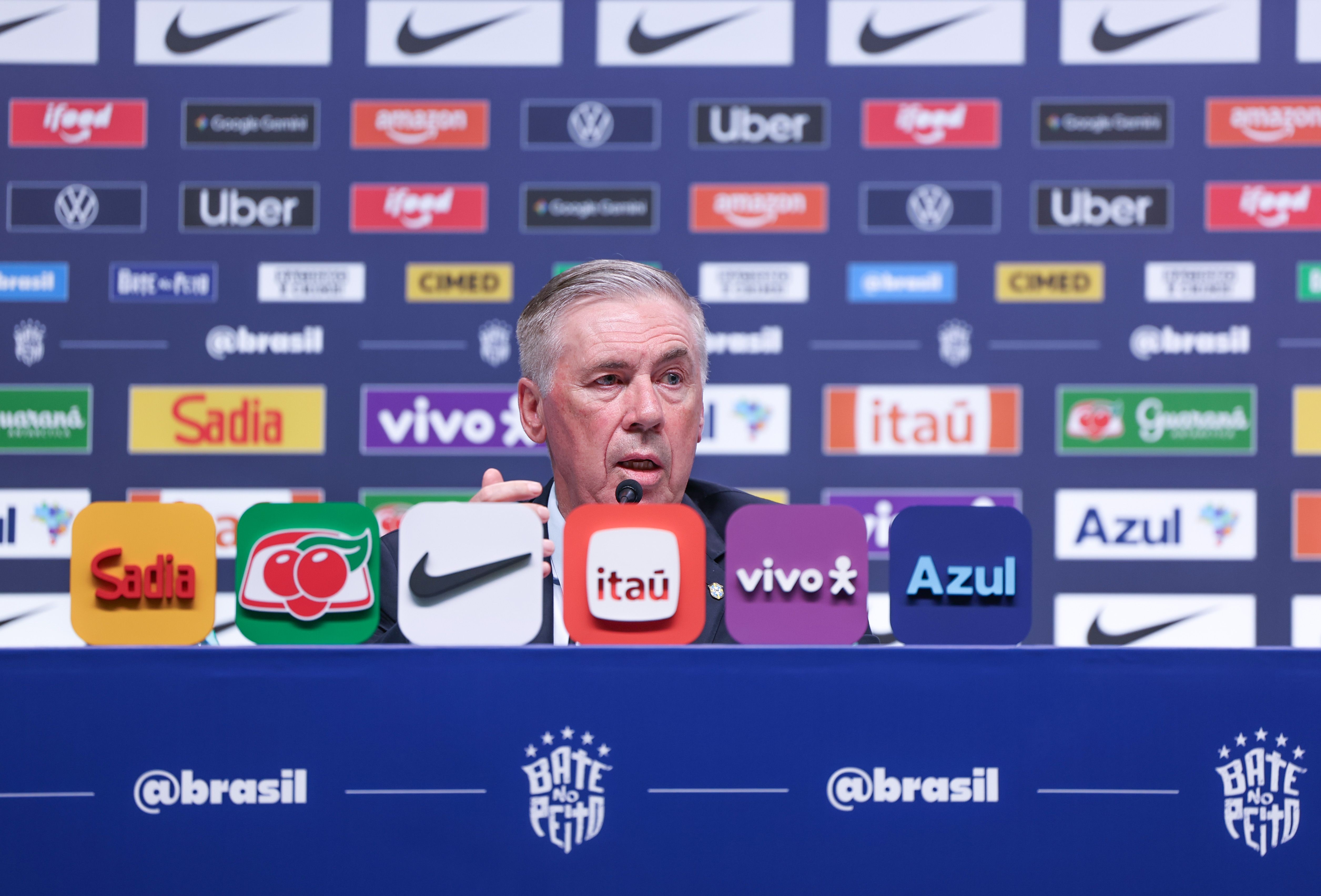 Homem de cabelos grisalhos, terno escuro e camisa branca, fala em um microfone durante coletiva de imprensa. À frente, logotipos de patrocinadores como Sadia, Guaraná, Nike, Itaú, Vivo e Azul. Ao fundo, um painel azul com diversos logotipos de marcas como Nike, Ifood, Amazon, Uber, Google Games, Cimed, Sadia, Vivo, Itaú e Guaraná. Na mesa, o texto @brasil e Bate no Peito