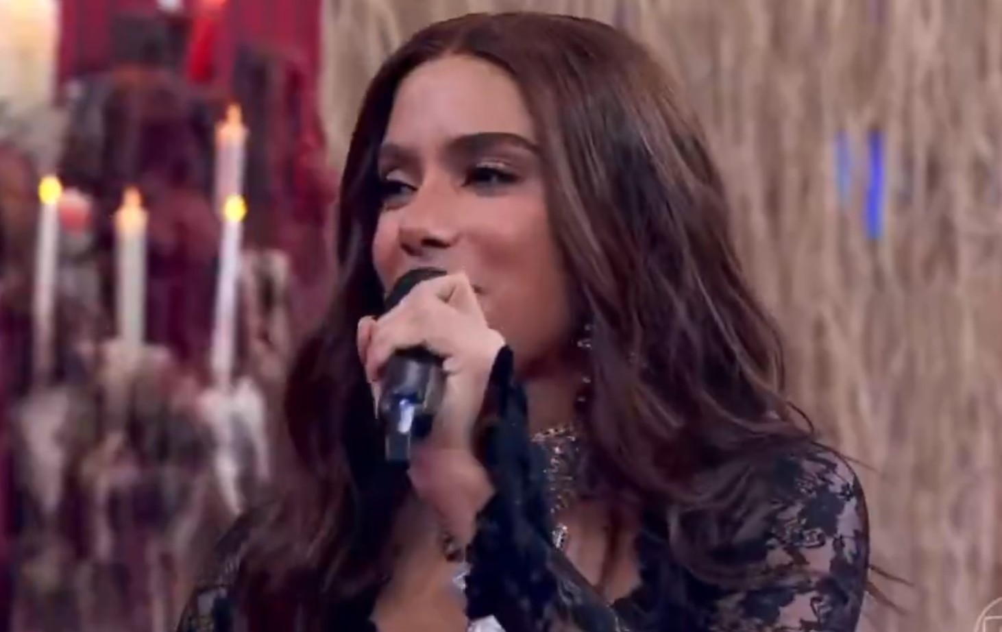 Cantora Anitta, com cabelos castanhos longos e maquiagem escura, sorri enquanto canta em um microfone preto. Ela veste uma blusa preta de renda, e ao fundo, velas acesas criam um ambiente intimista