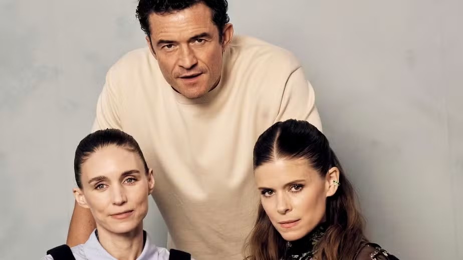 Três pessoas posam para foto: um homem de camiseta bege atrás, com duas mulheres à frente. A da esquerda tem cabelo preso e blusa clara, a da direita tem cabelo solto e blusa escura