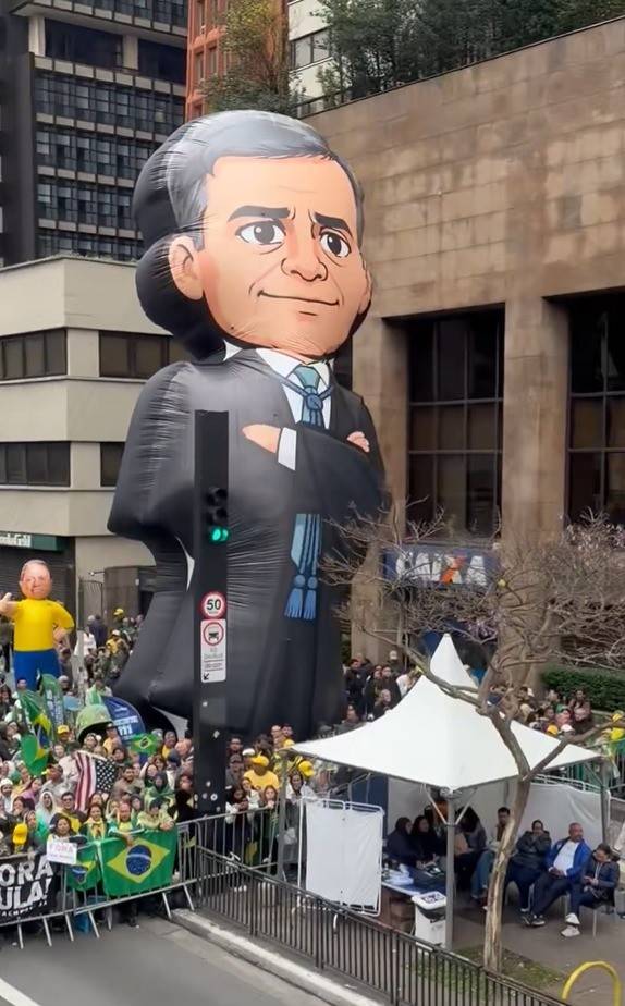 Bal&atilde;o gigante de um homem de terno preto e gravata azul, com bra&ccedil;os cruzados, em meio a uma multid&atilde;o de manifestantes segurando bandeiras do Brasil e cartazes, em uma rua urbana com pr&eacute;dios ao fundo