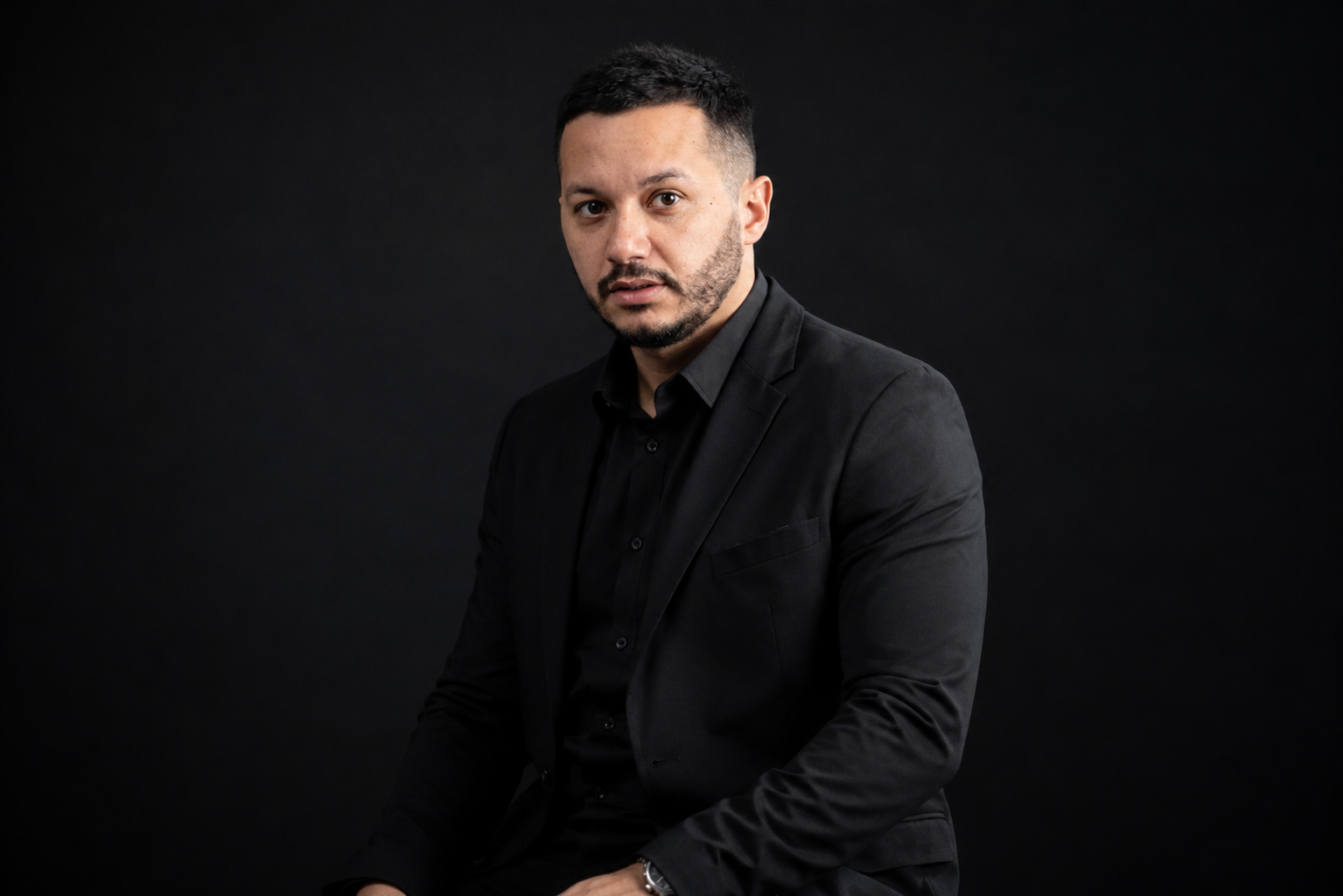 Homem de pele parda, barba e cabelo escuros, vestindo terno e camisa pretos, sentado e olhando diretamente para frente com expressão séria, contra um fundo preto