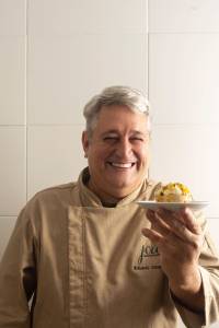Homem de cabelos grisalhos, vestindo d&oacute;lm&atilde; bege com o logo Joa Gastronomia e nome Eduardo Zam, sorri enquanto segura um pequeno bolo de maracuj&aacute; em um prato branco