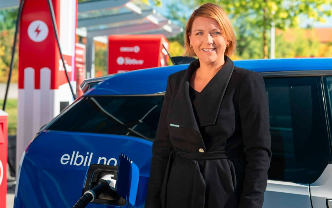 Mulher de cabelo ruivo e casaco preto sorri, em pé ao lado de um carro elétrico azul sendo carregado em um posto vermelho e branco