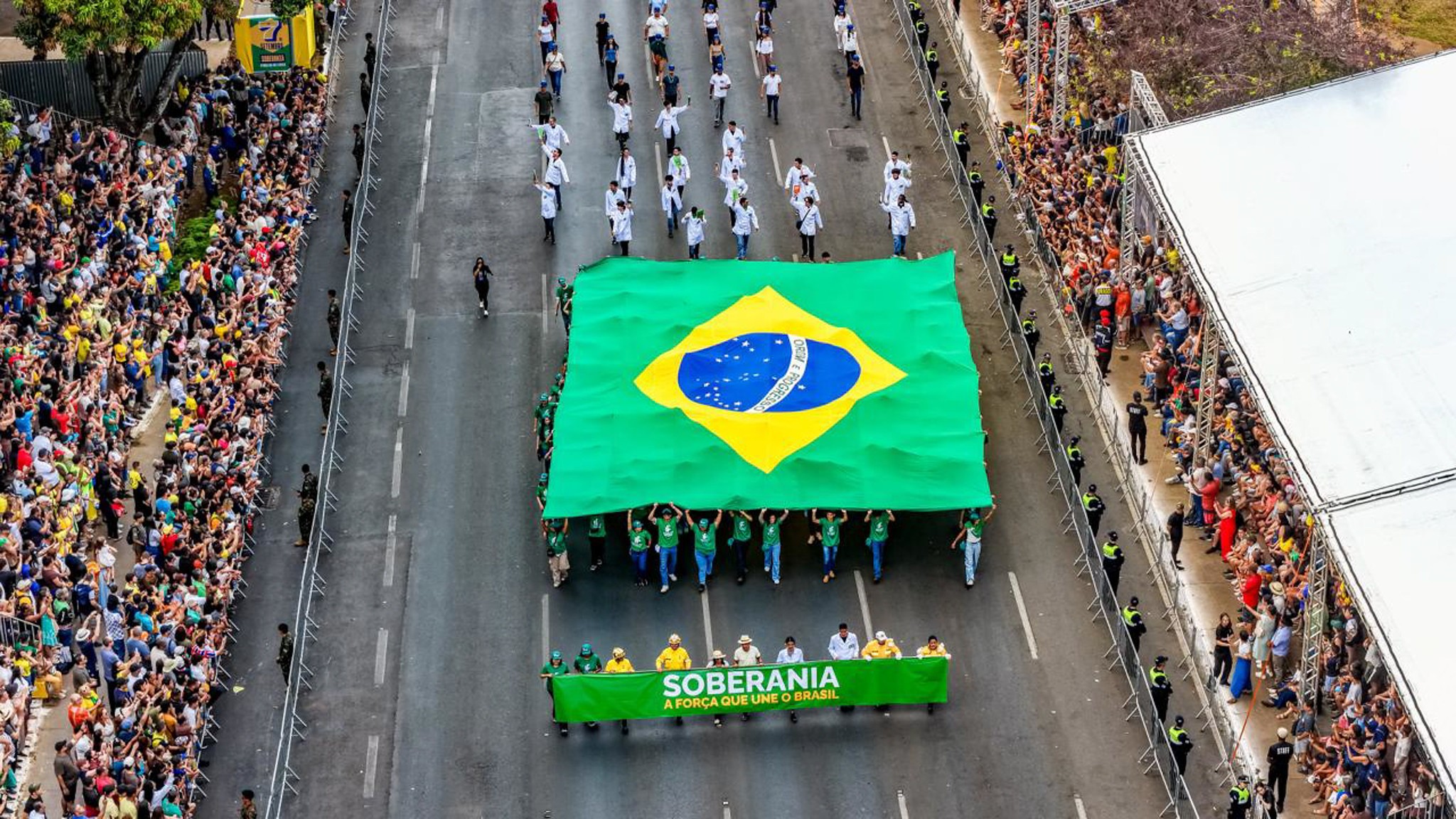 Vista aérea de uma multidão assistindo a um desfile cívico. No centro, pessoas carregam uma bandeira gigante do Brasil e uma faixa verde com a frase SOBERANIA A FORÇA QUE UNE O BRASIL.