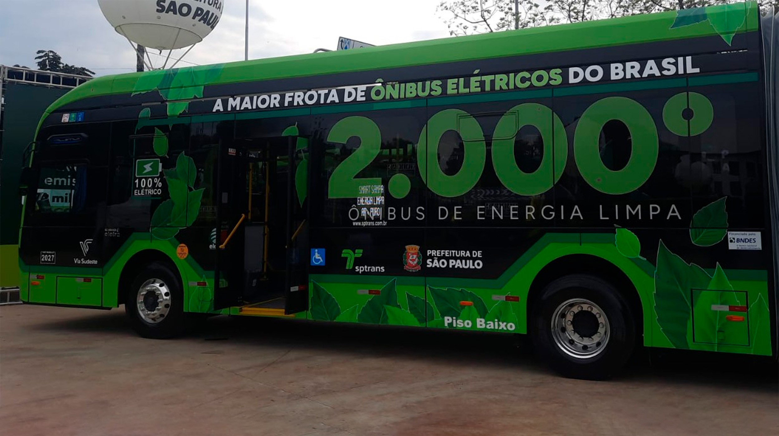 Ônibus elétrico preto e verde com a frase A MAIOR FROTA DE ÔNIBUS ELÉTRICOS DO BRASIL e o número 2.000 em destaque, indicando ÔNIBUS DE ENERGIA LIMPA. Há logos da SPTrans e Prefeitura de São Paulo, além de um balão branco ao fundo