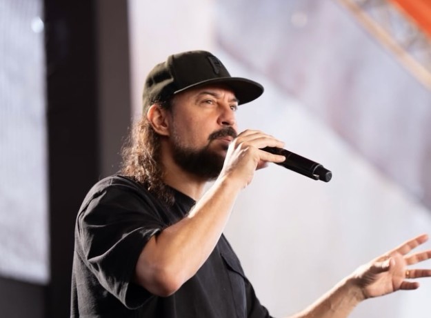 Homem de barba e cabelos longos e cacheados, usando bon&eacute; e camiseta preta, canta em um microfone preto enquanto gesticula com a m&atilde;o direita, em um palco iluminado