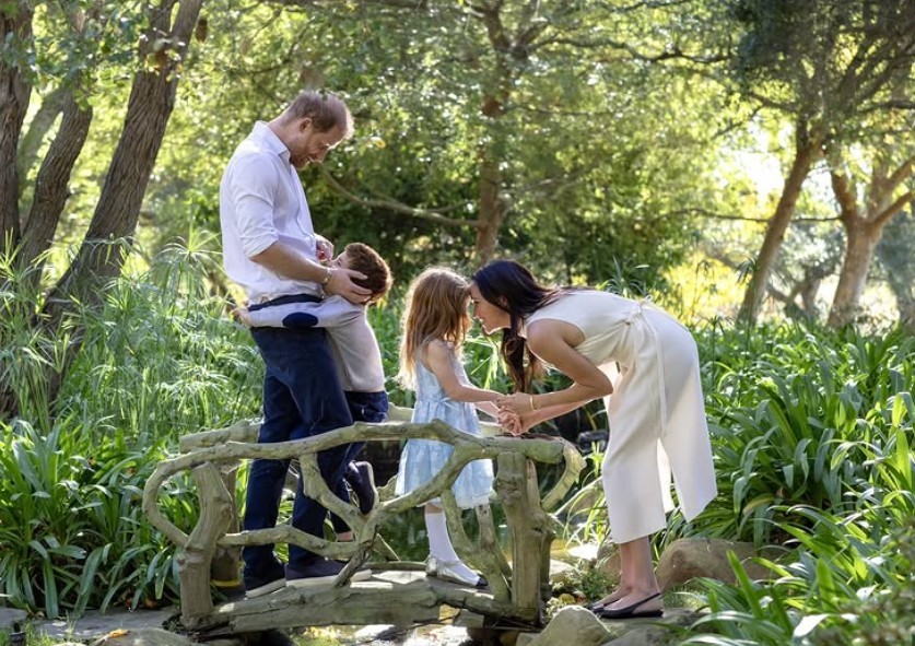 Família real em um jardim. O Príncipe Harry, de camisa branca e calça escura, abraça o filho Archie, de camisa clara. Meghan Markle, de vestido branco, inclina-se para beijar a filha Lilibet, de vestido azul claro. Todos estão sobre uma ponte rústica de madeira, cercados por vegetação exuberante e árvores ao fundo