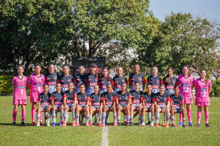 equipe de futebol feminino com 20 jogadoras, algumas em pé e outras sentadas em um banco de madeira, no gramado de um campo. Elas vestem uniformes de manga curta, pretos com detalhes vermelhos ou rosa choque, e meias esportivas. Ao fundo, árvores e céu azul claro
