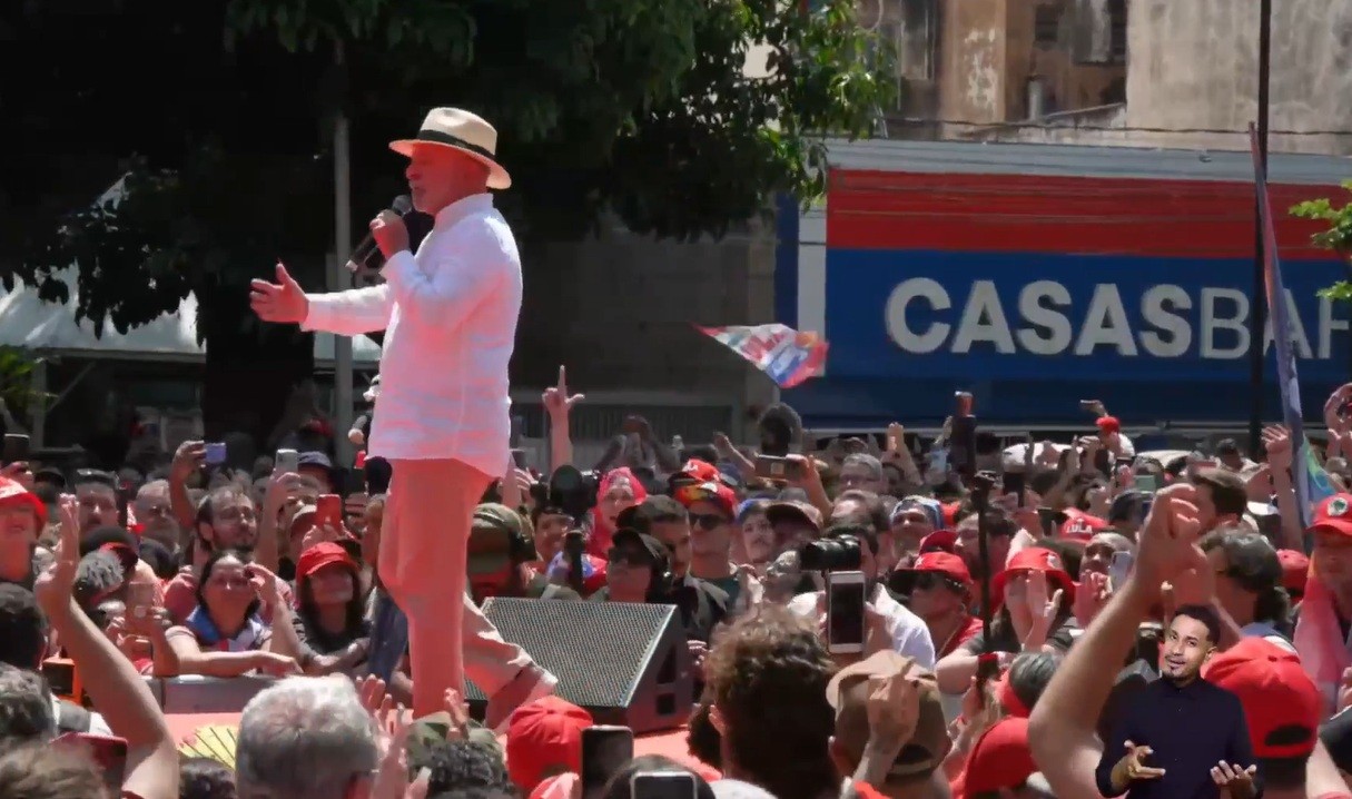 Homem de chapéu e camisa branca discursa em palco vermelho para multidão, com braços abertos e microfone. Pessoas com bonés vermelhos e celulares erguidos assistem. Ao fundo, fachada azul de loja Casas Bahia