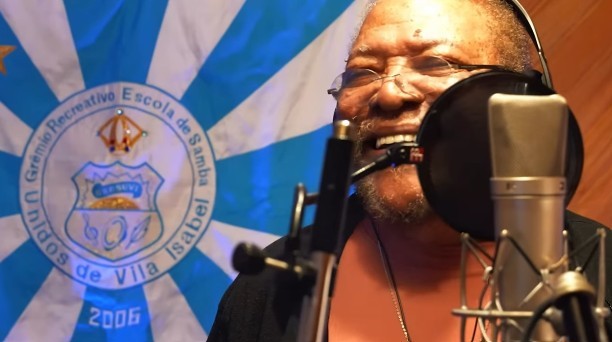 Homem negro de barba e óculos, usando fones de ouvido, canta em um microfone com filtro pop, sorrindo. Ao fundo, a bandeira azul e branca da Unidos de Vila Isabel, com o brasão da escola e o ano 2006