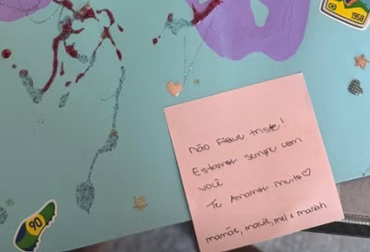 Um bilhete rosa escrito à mão com a mensagem Não fique triste! Estaremos sempre com você. Te amamos muito. Mamãe, Maui, Mel e Mariah, sobre uma superfície azul decorada com glitter vermelho e roxo, e adesivos de coração, estrela e chuteira