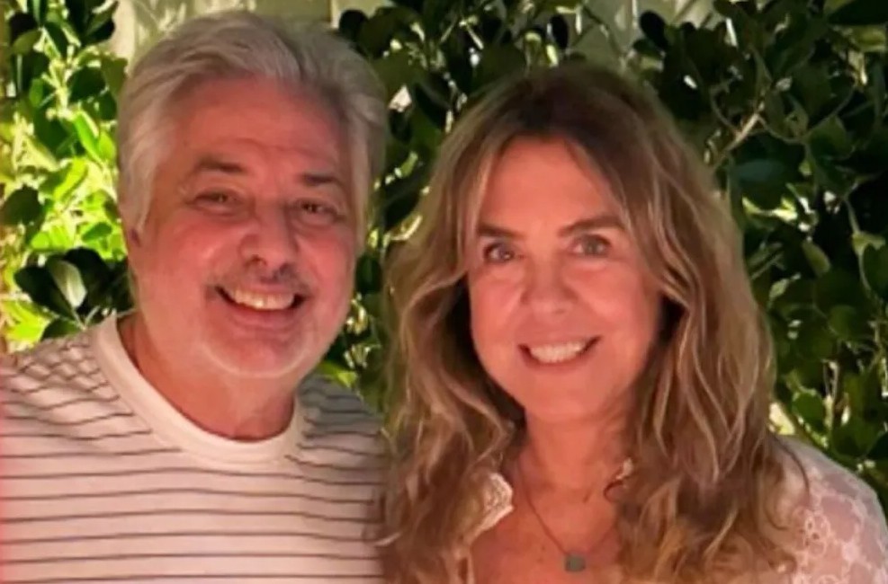 Um homem de cabelos grisalhos e uma mulher de cabelos loiros, ambos sorrindo, posam para a câmera com folhagens verdes ao fundo