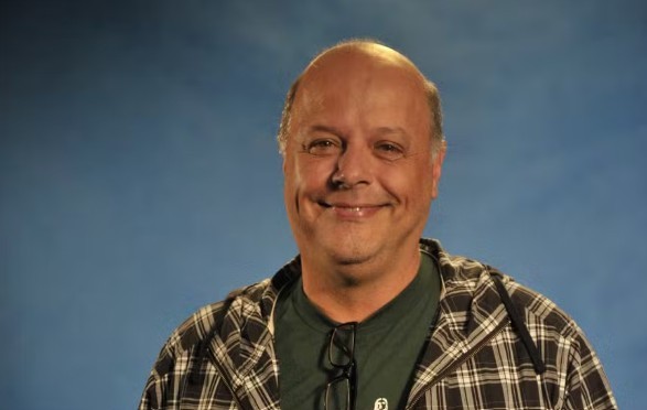 Homem calvo de meia-idade, sorrindo, usando camisa xadrez preta e branca sobre camiseta verde, com óculos pendurados na gola, em fundo azul