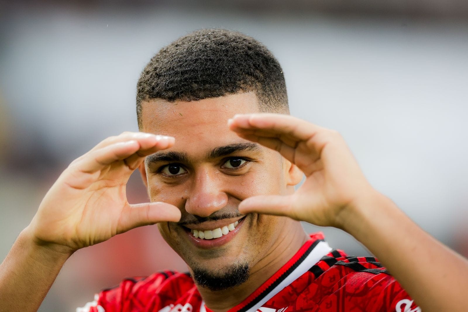Homem negro de cabelo curto e barba rala, sorrindo e fazendo um coração com as mãos em frente ao rosto, vestindo camisa de futebol vermelha e preta