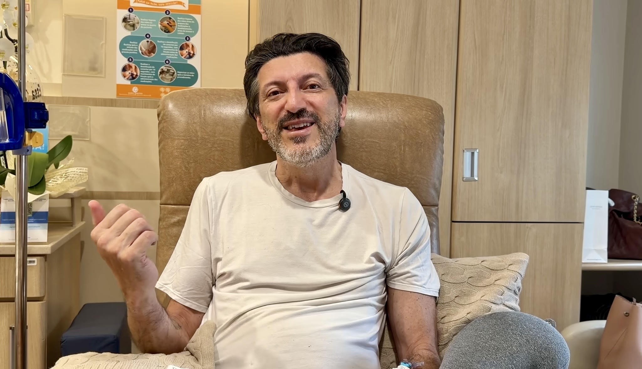 Homem de meia-idade, cabelo escuro e barba grisalha, sorri para a câmera enquanto está sentado em uma poltrona marrom. Ele veste uma camiseta bege e faz um sinal de positivo com a mão esquerda. Ao fundo, um armário de madeira clara e um suporte de soro.