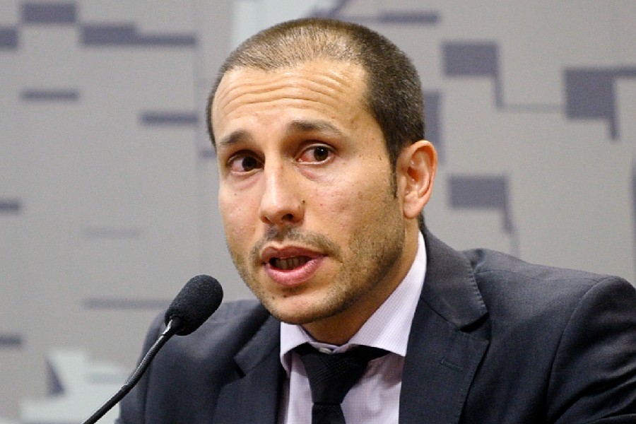 Homem de pele clara, cabelo raspado e barba rala, vestindo terno escuro e gravata preta, fala em um microfone preto. Ele tem a boca ligeiramente aberta e os olhos arregalados, olhando para a direita. O fundo é cinza com formas geométricas claras