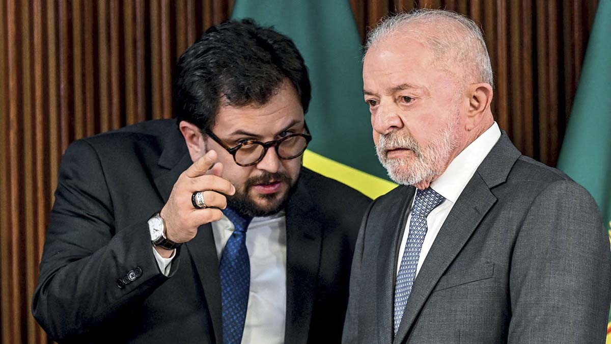 Dois homens em trajes sociais. À esquerda, um homem de barba e óculos aponta o dedo indicador para frente, falando com o homem à direita, que tem barba branca e expressão séria. Ao fundo, bandeiras do Brasil.