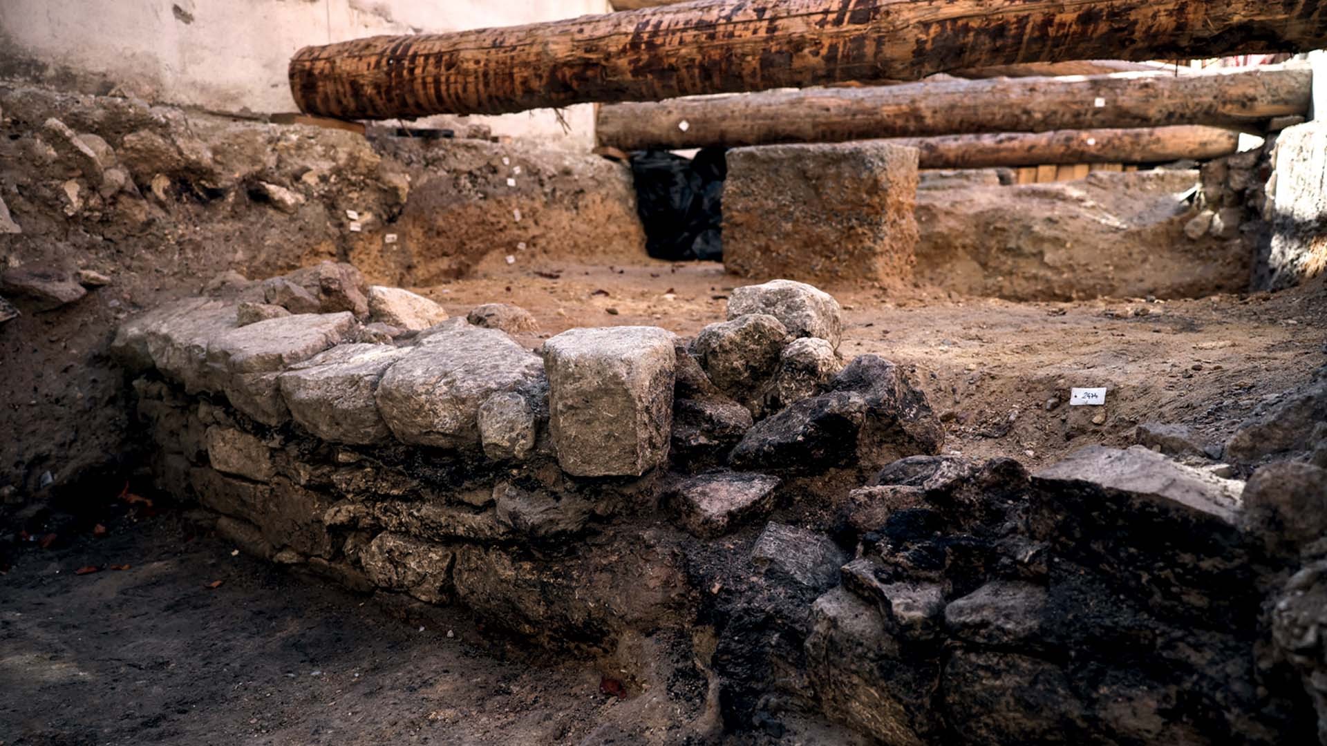 Sítio arqueológico com paredes de pedra e terra expostas, revelando camadas de solo e estruturas antigas. Dois troncos de madeira grossos e escuros atravessam a parte superior da imagem, apoiados sobre blocos de pedra e terra. O chão é de terra batida, com algumas pedras soltas e um pequeno pedaço de papel branco no centro. A iluminação lateral cria sombras e realça a textura dos materiais.