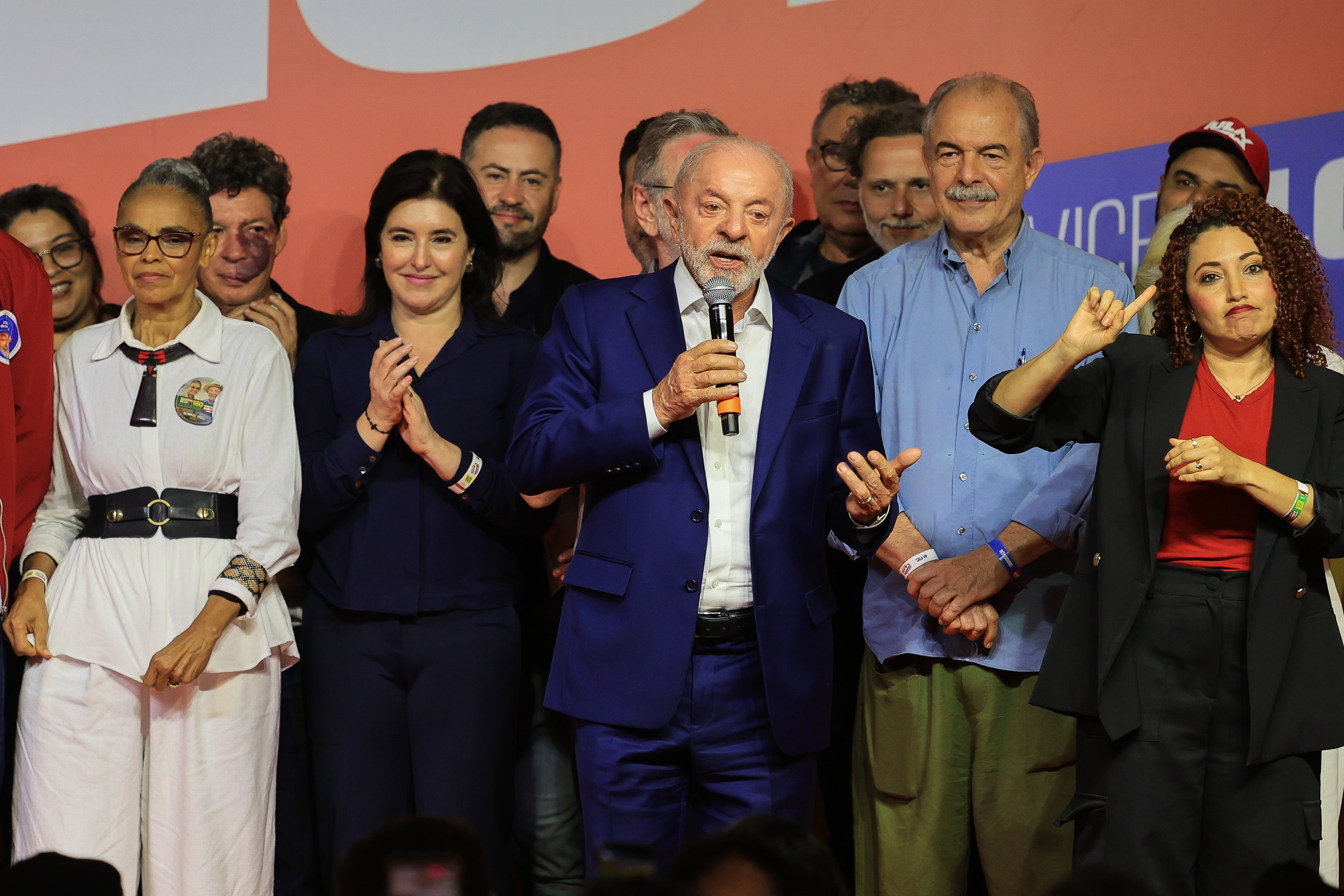 Lula, de terno azul, fala ao microfone em um palco com outras pessoas. À esquerda, uma mulher de roupa branca e óculos, e outra de blusa azul escura aplaudindo. À direita, uma mulher de blusa vermelha e blazer preto faz um gesto com a mão. Outros homens observam ao fundo