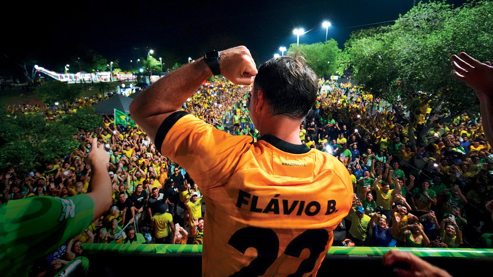 Homem de costas, vestindo camisa amarela com FLÁVIO B e 22 nas costas, braço direito levantado com punho cerrado, saudando uma multidão vibrante em evento noturno. Pessoas vestem roupas amarelas e verdes, algumas seguram bandeiras e celulares acesos, sob iluminação de postes e árvores.