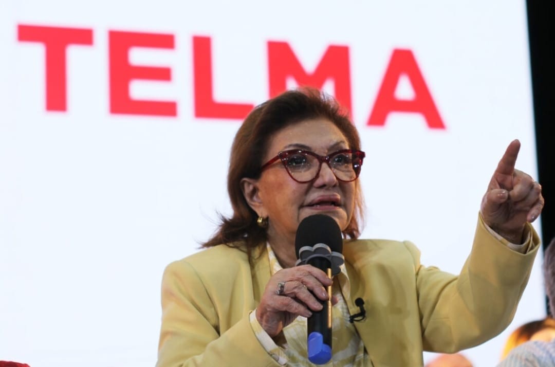 Mulher de cabelos castanhos e óculos vermelhos, vestindo blazer amarelo, fala ao microfone e aponta o dedo indicador para cima, com a palavra TELMA em vermelho ao fundo