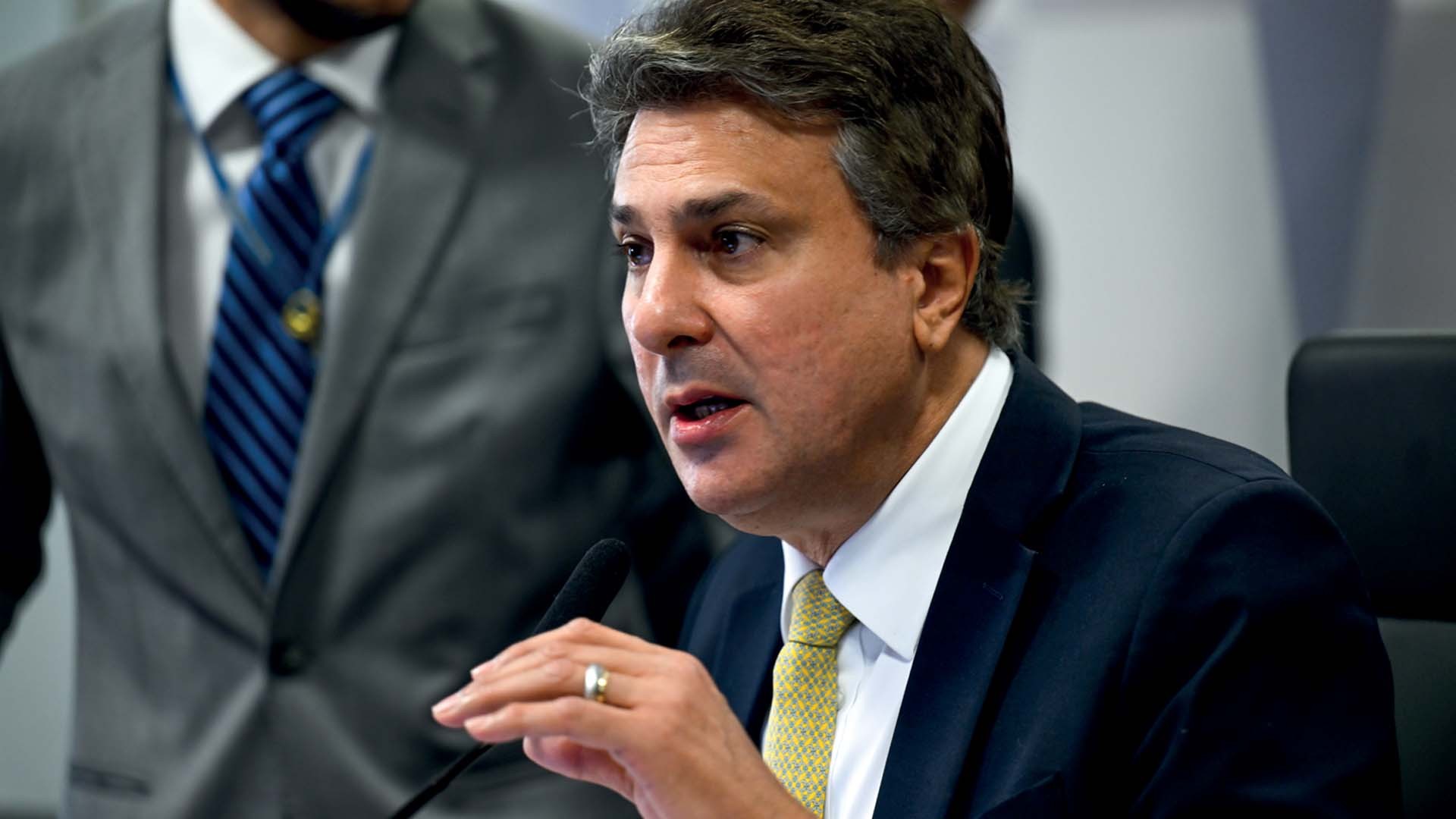Homem de meia-idade, cabelos escuros e ondulados, terno azul-marinho, camisa branca e gravata amarela, falando em um microfone. Ele usa um anel dourado na mão direita. Ao fundo, outro homem de terno cinza e gravata azul listrada está desfocado.