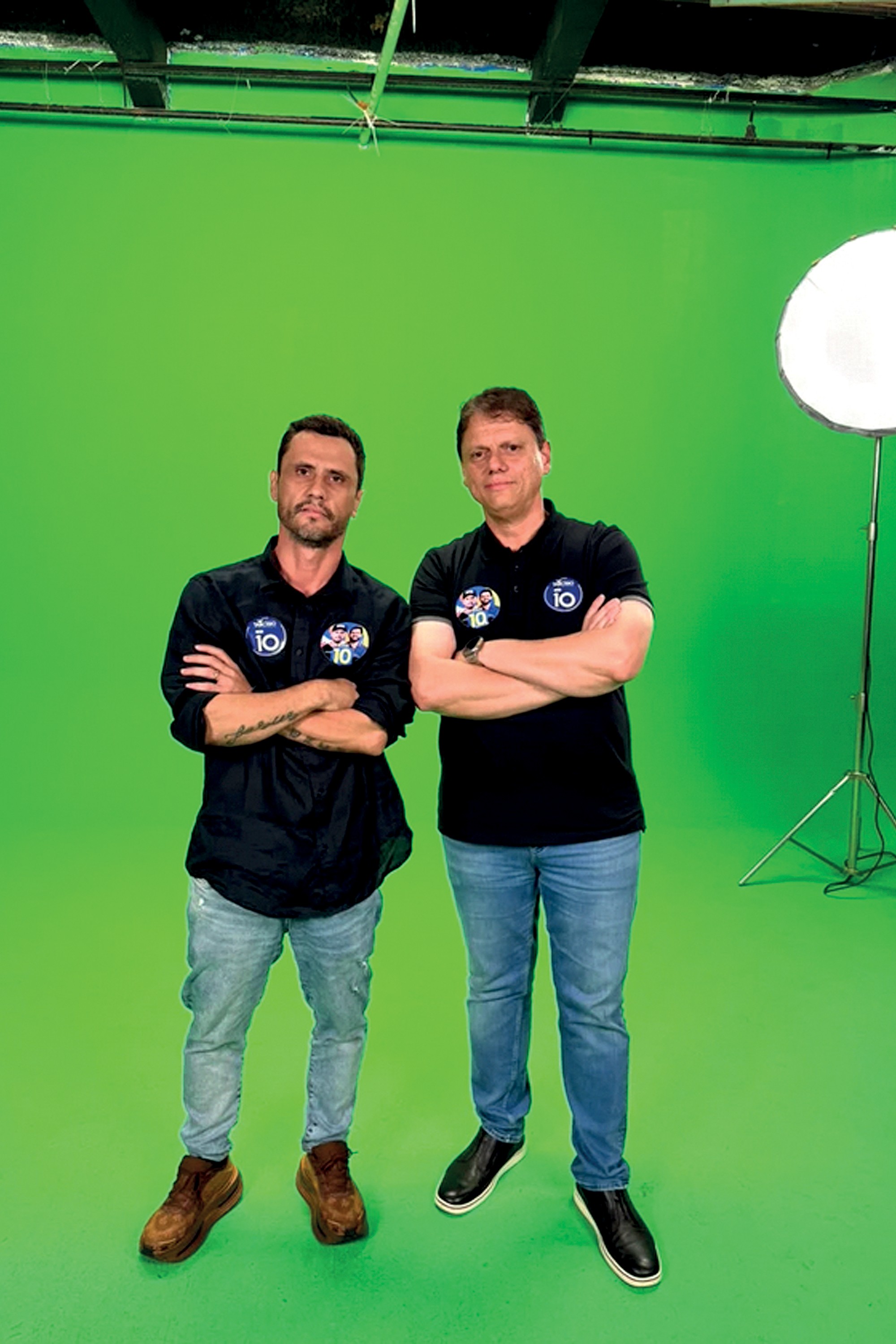 Dois homens, um barbudo e outro de cabelo curto, vestem camisas pretas com broches 10 e calças jeans, posando de braços cruzados em um estúdio com fundo verde e um softbox à direita