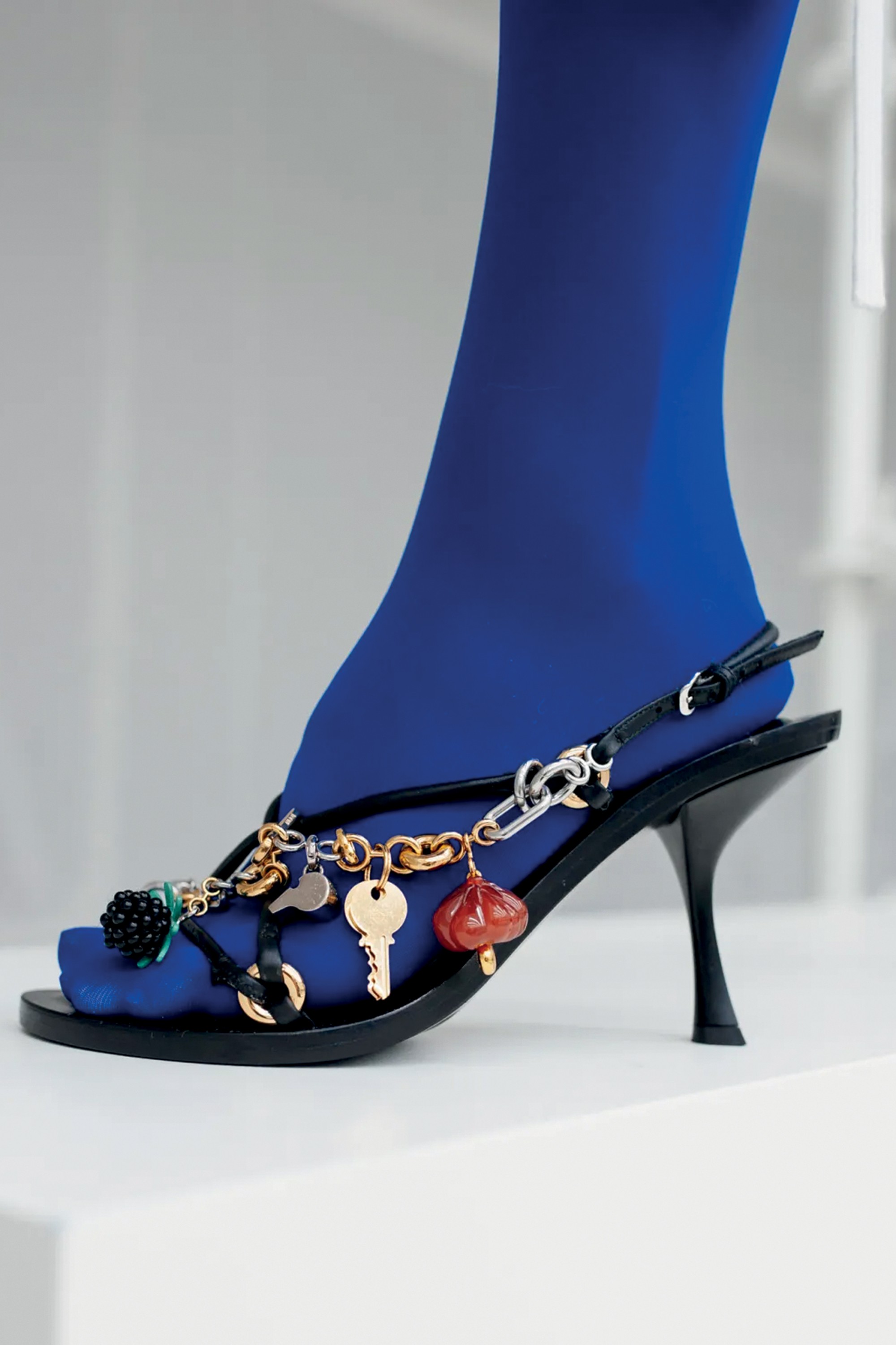 Perna com meia-calça azul royal e sandália preta de salto fino, decorada com correntes douradas e prateadas, chaves, um pingente de amora preta e outro vermelho em formato de cogumelo