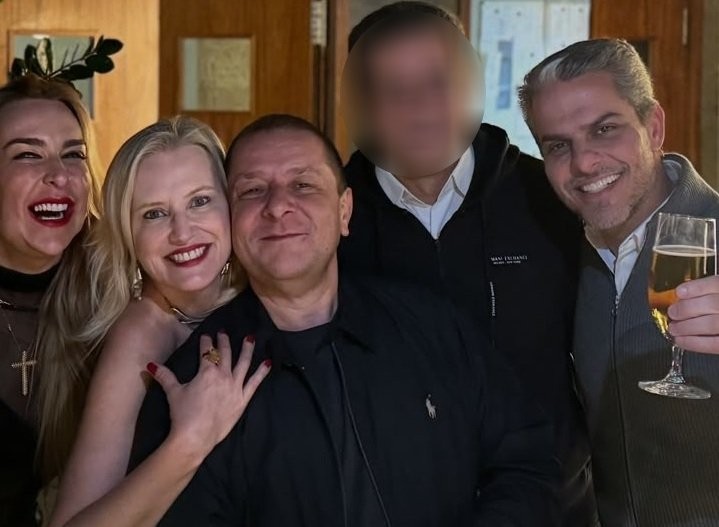 Cinco pessoas sorrindo para a câmera. Uma mulher loira abraça um homem de camisa preta. À esquerda, outra mulher loira ri, usando tiara de folhas. À direita, um homem de cabelo grisalho segura uma taça de champanhe. Um quinto homem tem o rosto borrado